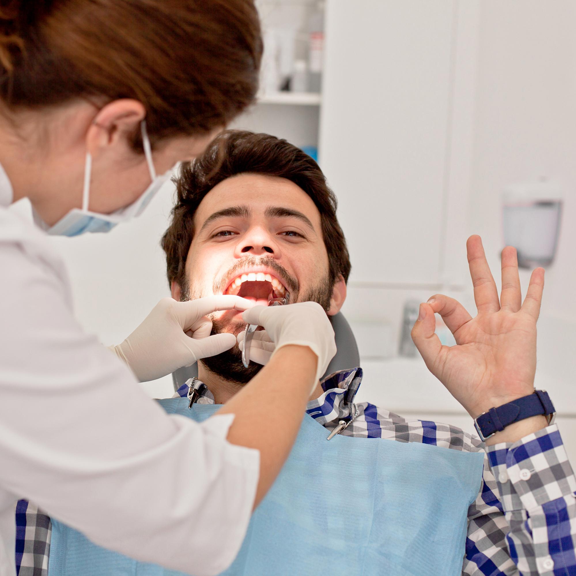 man smiling at the dentist with his mouth open whilst sticking up his hand and showing an okay sign to the camera